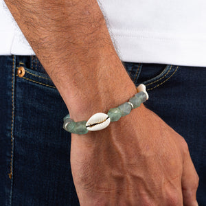 Person wearing a bracelet with green beads and a cowrie shell on a white shirt and blue jeans background.