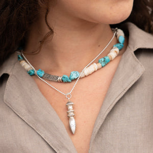 Necklace with turquoise and white beads and a silver pendant worn by a person.
